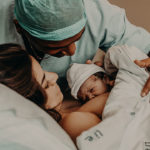 mom in hospital bed after c-section with newborn baby during skin-to-skin time and father leaning over them